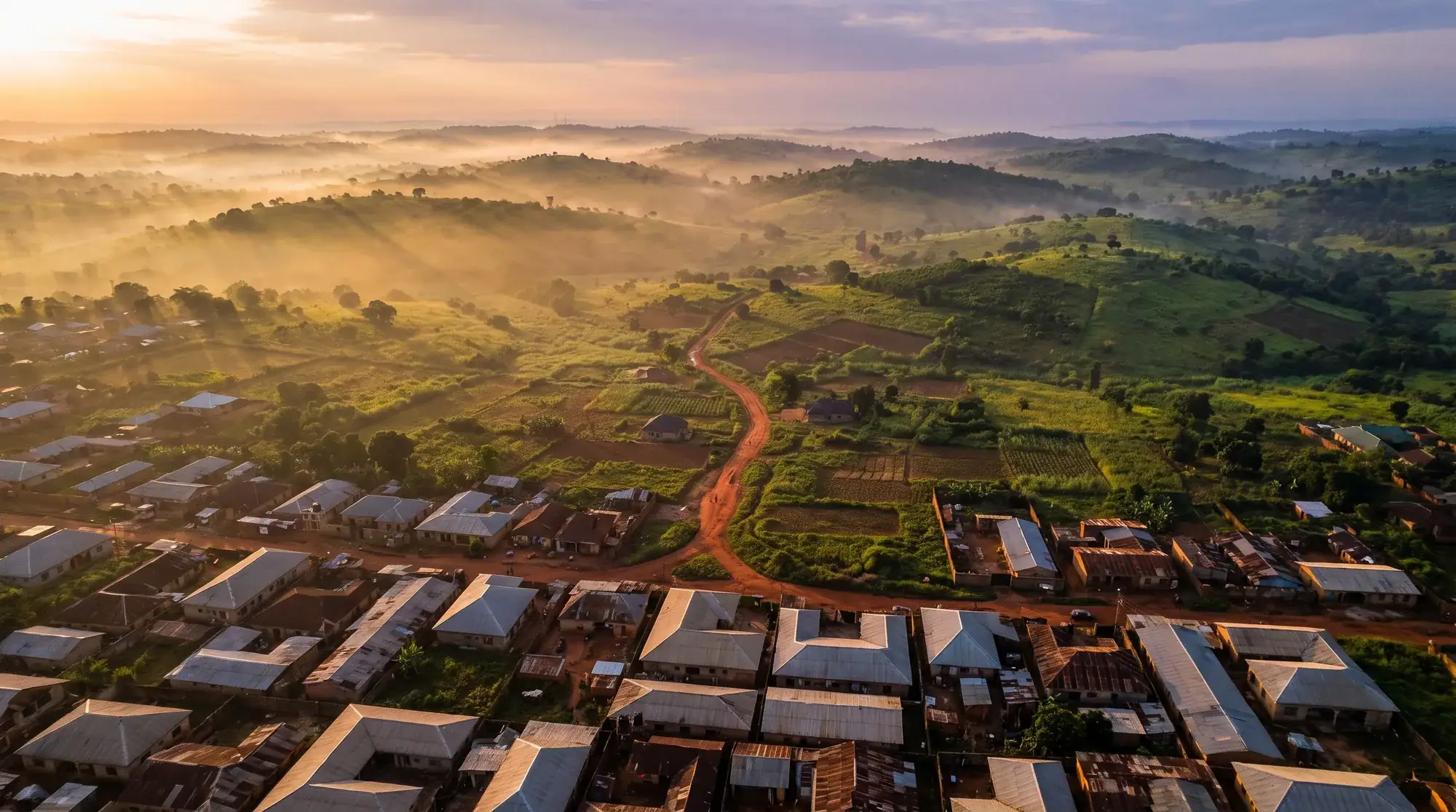 An aerial view of a Nigerian city edge dissolving into green countryside at dawn.